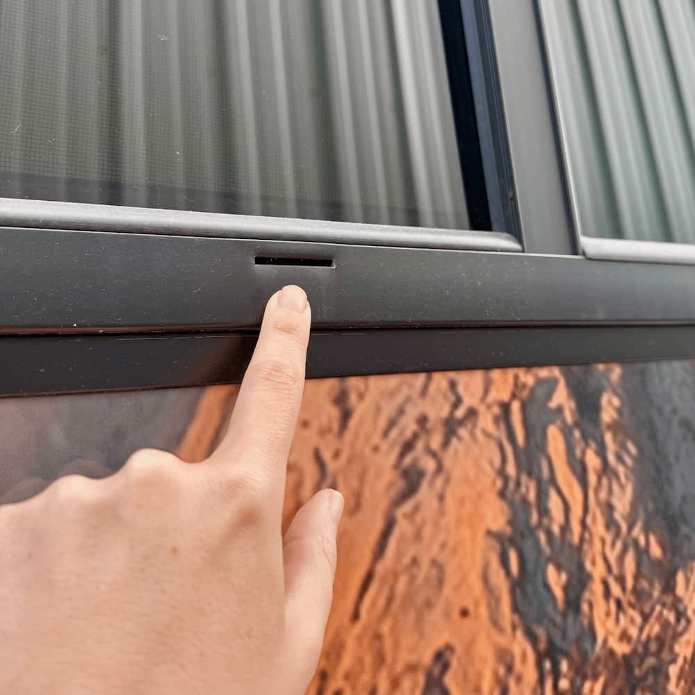 A person is pointing a finger at the opening on the window trim to indicate the weep hole.